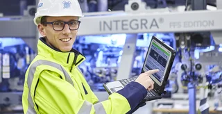 Ein Mitarbeiter mit Schutzhelm, Brille und gelber Sicherheitsjacke demonstriert Leistungsdaten auf einem Laptop.