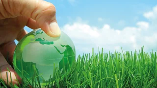 Hand holds green glass sphere with world map over grass, symbol of sustainable energy and environmental awareness under blue sky with clouds.