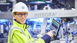An employee wearing a hard hat, goggles and yellow safety jacket demonstrating performance data on a laptop.