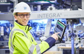 Employee wearing a hard hat, goggles and yellow safety jacket demonstrating performance data on a laptop.