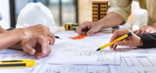 Hands of professionals over construction plans during an architectural project meeting, with tools and hard hats on the table.