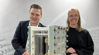 Lars Wegner und Christina Kemper von Haver & Boecker lächeln hinter einem technischen Filtermodul auf einem Holztisch, im Hintergrund Drahtgeflecht-Muster.