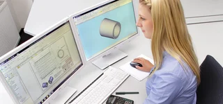 A young product designer works with CAD software on a 3D model and technical drawing of a metal component on two monitors in the design office.