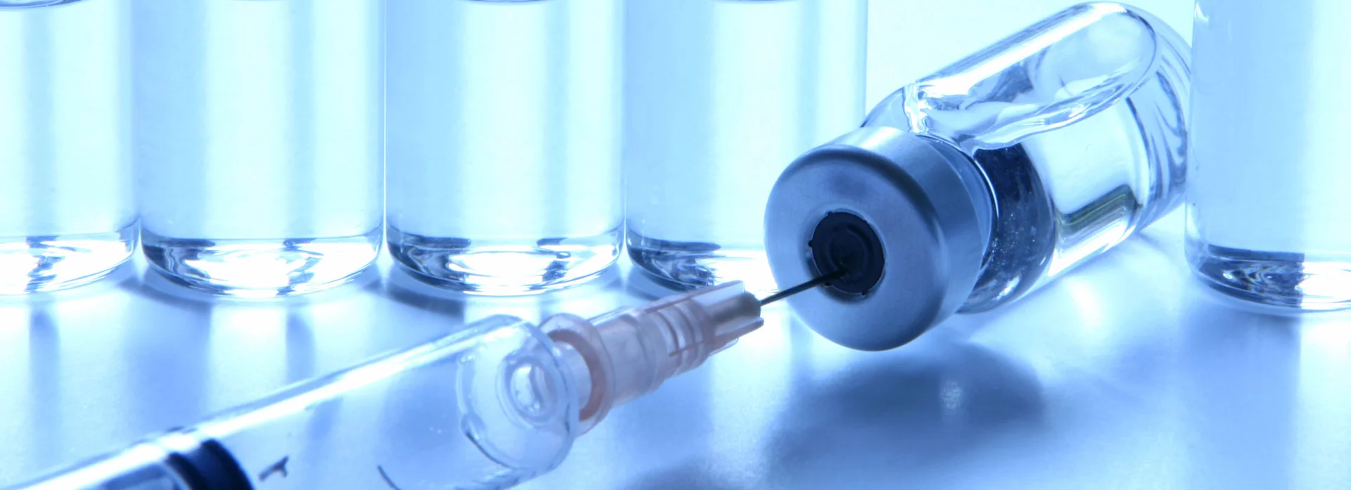 Medical glass ampoules containing clear liquid, in the foreground a syringe inserted into the rubber stopper of a reclining ampoule.