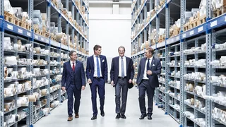 Vier Gesellschafter von Haver & Boecker in Anzügen gehen lächelnd durch ein Hochregallager, umgeben von gelagerten Drahtgeweberollen.