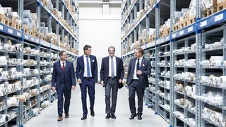Four shareholders of Haver & Boecker in suits walking through a high‑bay warehouse, smiling and surrounded by stored wire mesh rolls.