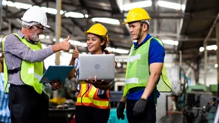 Three people in protective clothing standing at the laptop and giving a smiling thumbs-up.