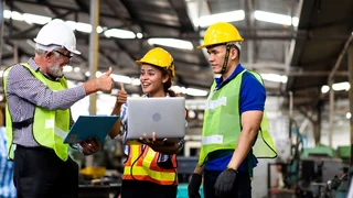 Drei Personen in Schutzbekleidung stehen an Laptop und strecken lächelnd die Daumen hoch.