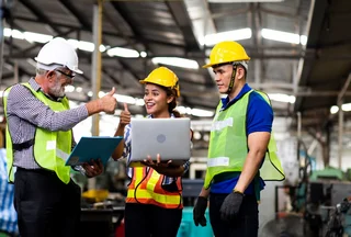 Three people in protective clothing standing at the laptop and giving a smiling thumbs-up.
