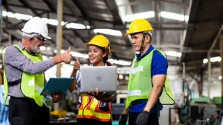 Drei Personen in Schutzbekleidung stehen an Laptop und strecken lächelnd die Daumen hoch.