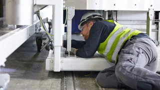 Ein Mann mit Schutzhelm und Warnweste montiert etwas unter einer Maschine liegend.