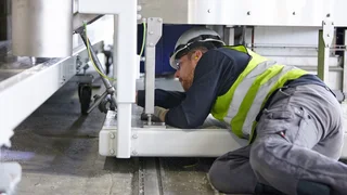 Man with safety helmet and high-visibility vest mounting something while lying under a machine.