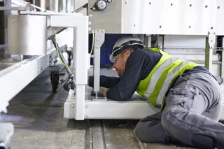 Man with safety helmet and high-visibility vest mounting something while lying under a machine.