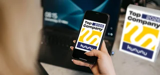 Hand holding smartphone with Kununu award “Top Company 2025”, in the background another Kununu sign and laptop on desk.