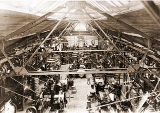 Historic weaving hall of Haver & Boecker in sepia, with numerous mechanical looms and steel beams under an open roof truss.